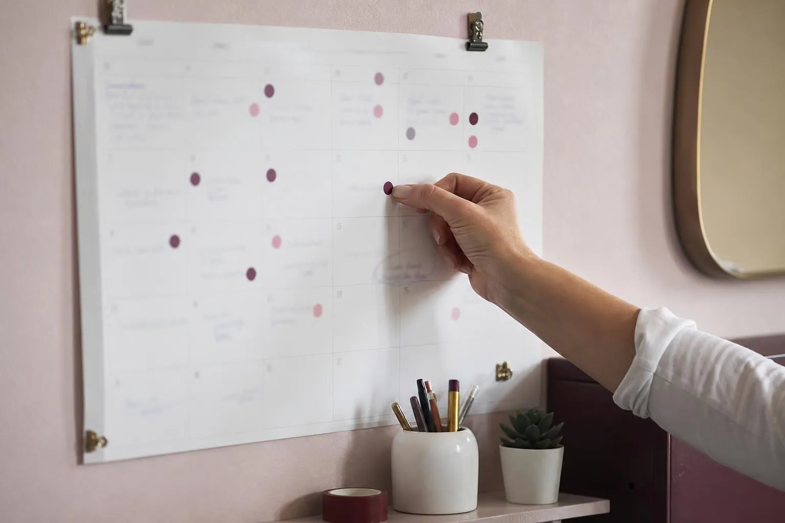 Main d’une praticienne collant une pastille de couleur sur un calendrier mural annoté, accroché au mur rose poudre d’une cabine