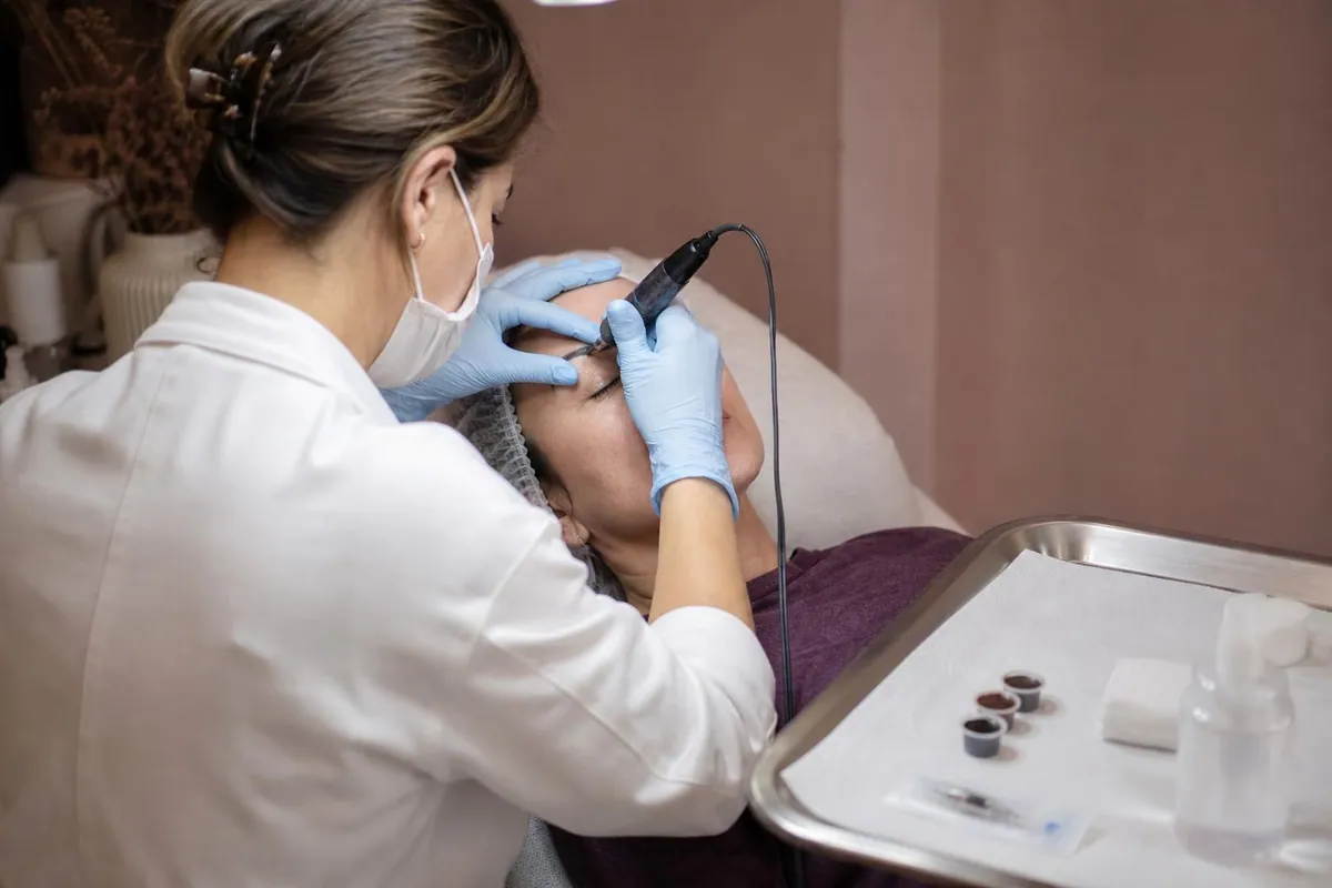 Mains gantées d’une praticienne tenant un dermographe au dessus du sourcil d’une cliente allongée, plateau inox avec godets de pigment et cartouche d’aiguilles à côté